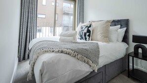 A bedroom with a bed, a bedside table, a window with curtains, and a view of buildings outside.