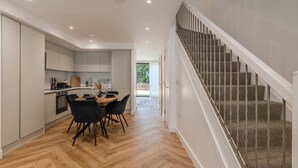 A modern kitchen with white cabinets, a dining area with a wooden table and black chairs, and a staircase with patterned carpet.