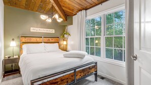Un dormitorio con techo de madera, un ventanal grande, una cama con sábanas blancas, una mesita de noche con una lámpara y un cartel en la pared.