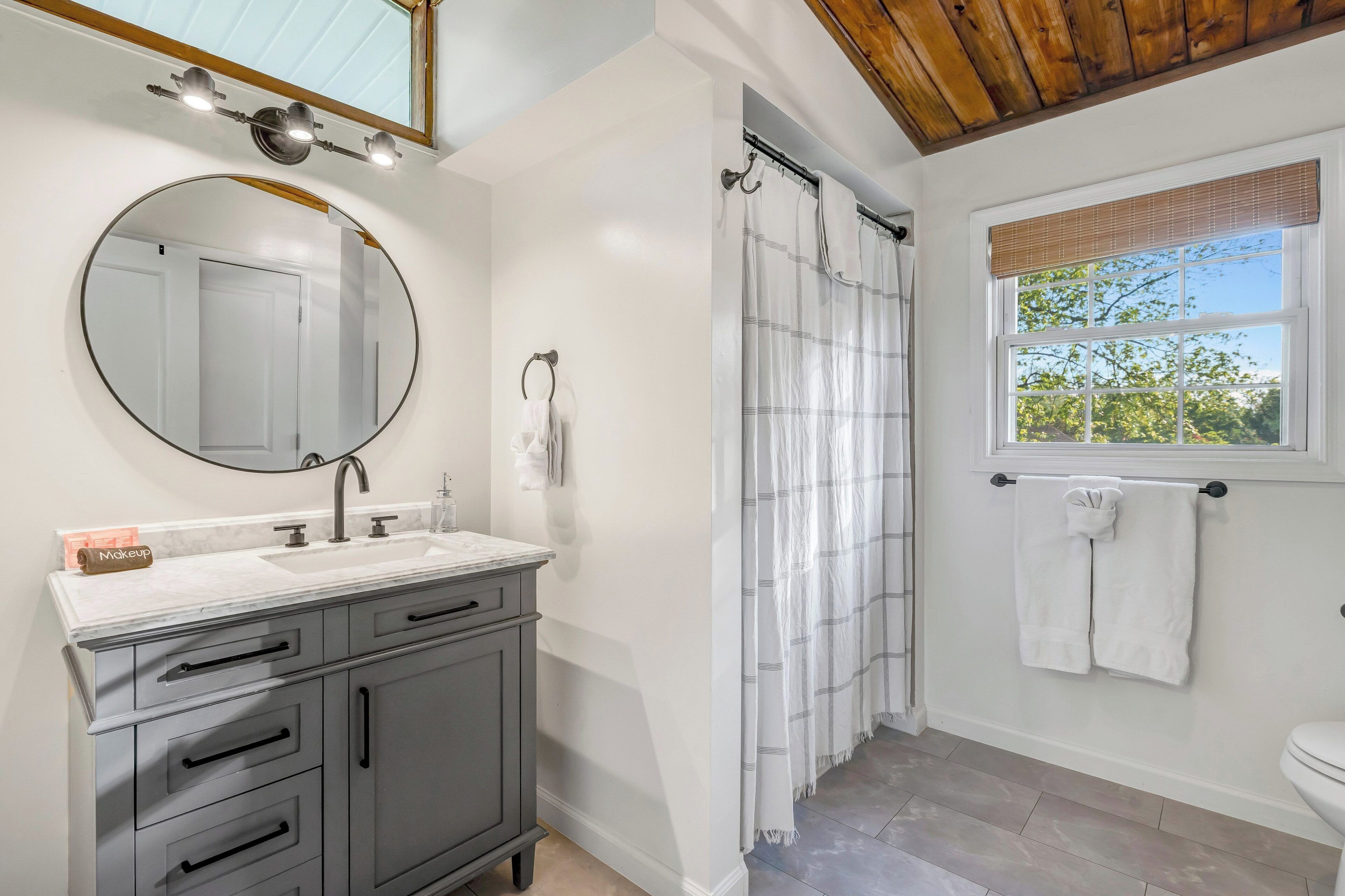 A bathroom with a round mirror, a vanity with drawers, a shower curtain, a window with a view, and a towel rack.