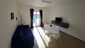 Una sala de estar moderna con un sofá azul, una mesa de comedor con sillas, un televisor sobre un mueble negro y un balcón con cortinas.