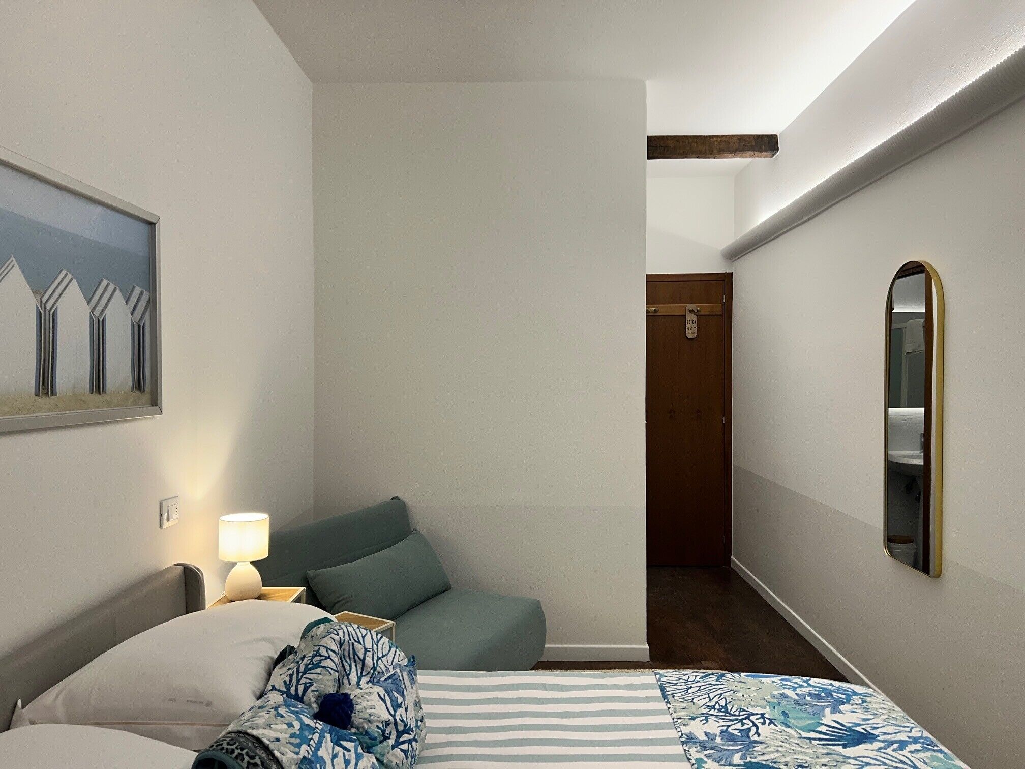 A hotel room with a bed, a bedside table with a lamp, a green armchair, a mirror, and a door leading to another room.