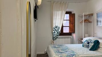 A bedroom with a bed, a wooden ceiling, a radiator, a window with curtains, a coat rack, and a wall-mounted light fixture.
