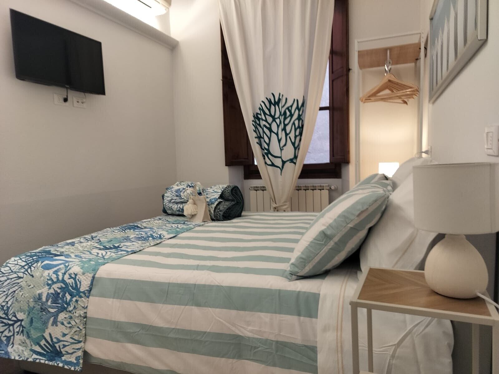 Un dormitorio con cama, televisor montado en la pared, mesita de noche con lámpara y ventana con cortinas.