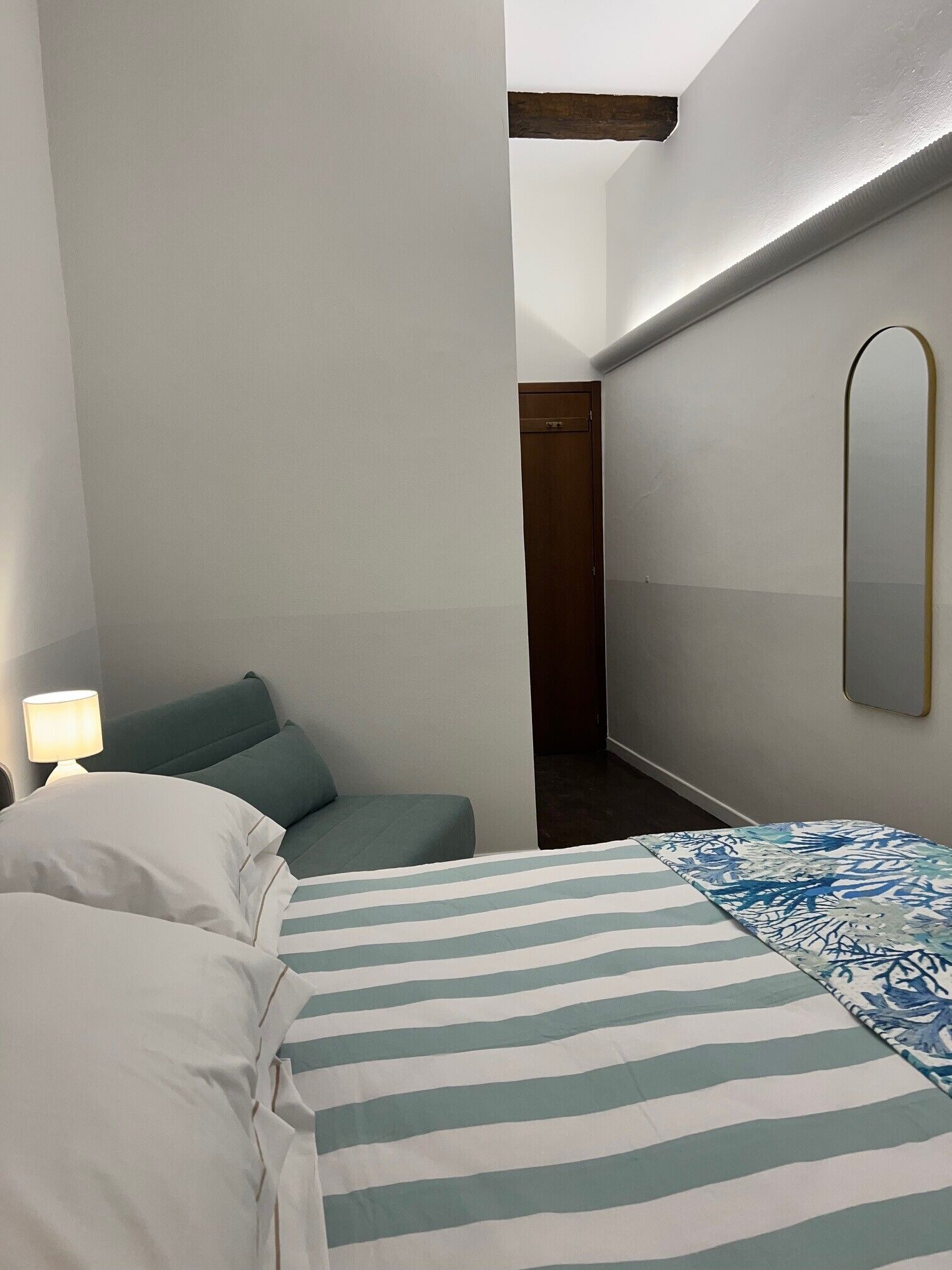 Hotellihuone, jossa on sänky, yöpöytälamppu, pieni oleskelualue ja ovi, joka johtaa toiseen huoneeseen.
