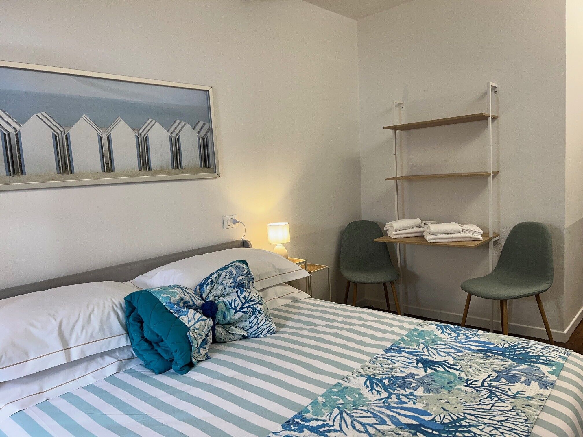 A hotel room with a bed, two chairs, a small table with towels, a wall-mounted shelf, and a painting.