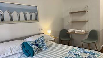 A hotel room with a bed, two chairs, a small table with towels, a wall-mounted shelf, and a painting.