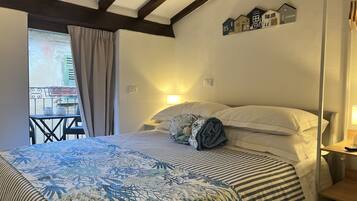 A bedroom with a bed, a nightstand, a window with curtains, a balcony with chairs, and a wall-mounted shelf with house-shaped decorations.