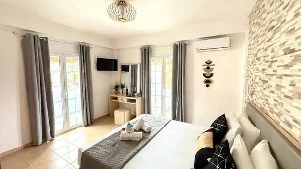 A modern hotel room with a large bed, a flat-screen TV, a desk with a chair, and a view of the outdoors through the windows. - Casa Damour  (Sidari)