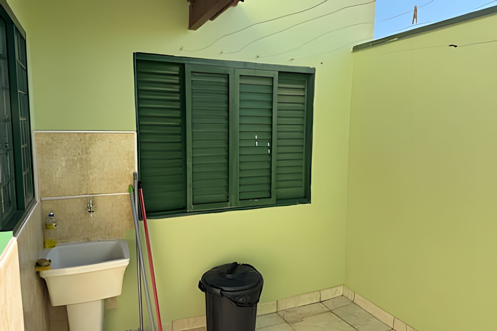 A small bathroom with a sink, a window with green shutters, and a trash bin.