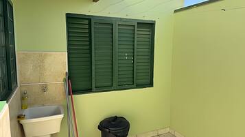 A small bathroom with a sink, a window with green shutters, and a trash bin.