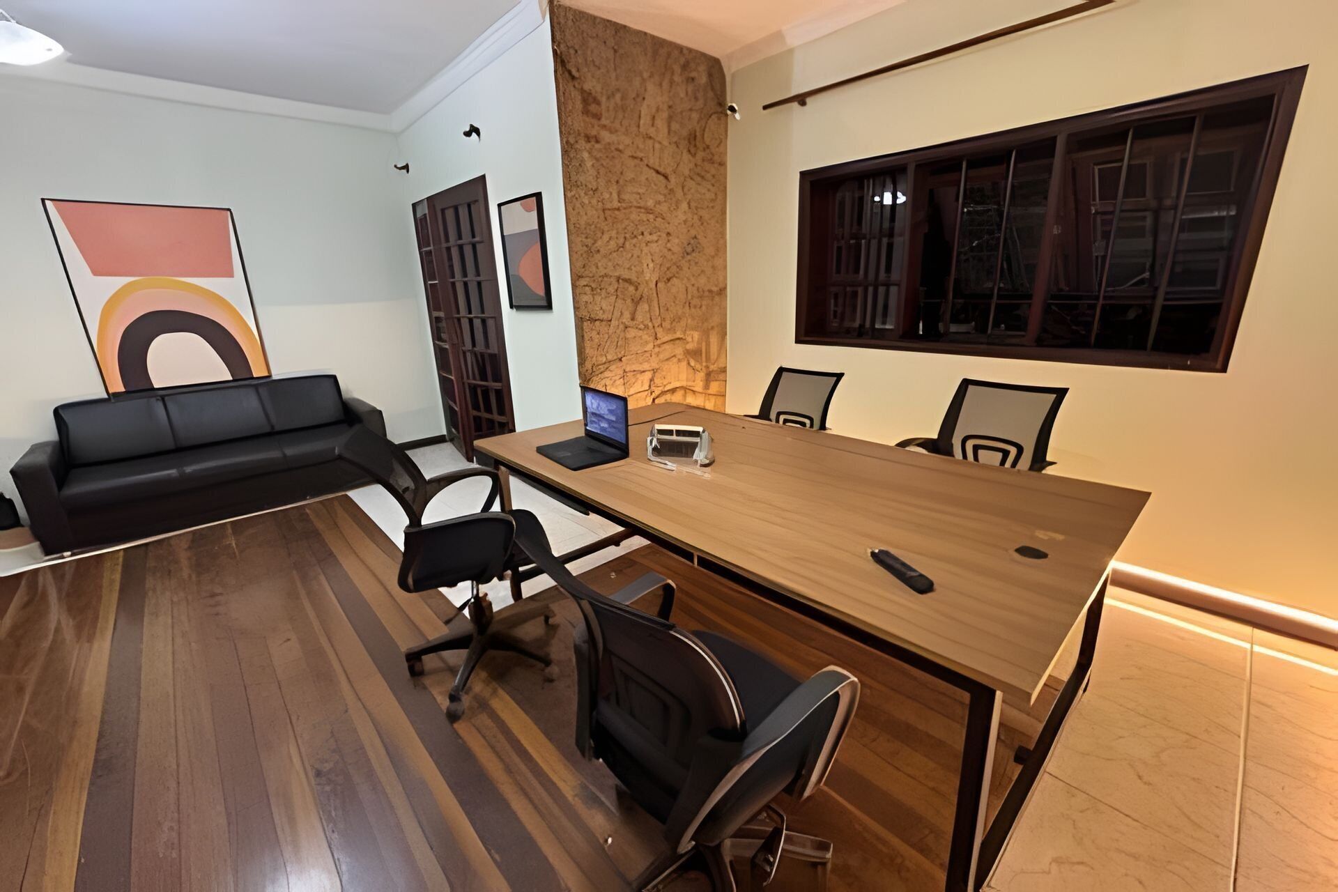 Un salón de conferencias con una mesa de madera, sillas negras, una computadora portátil y un gran ventanal con persianas.