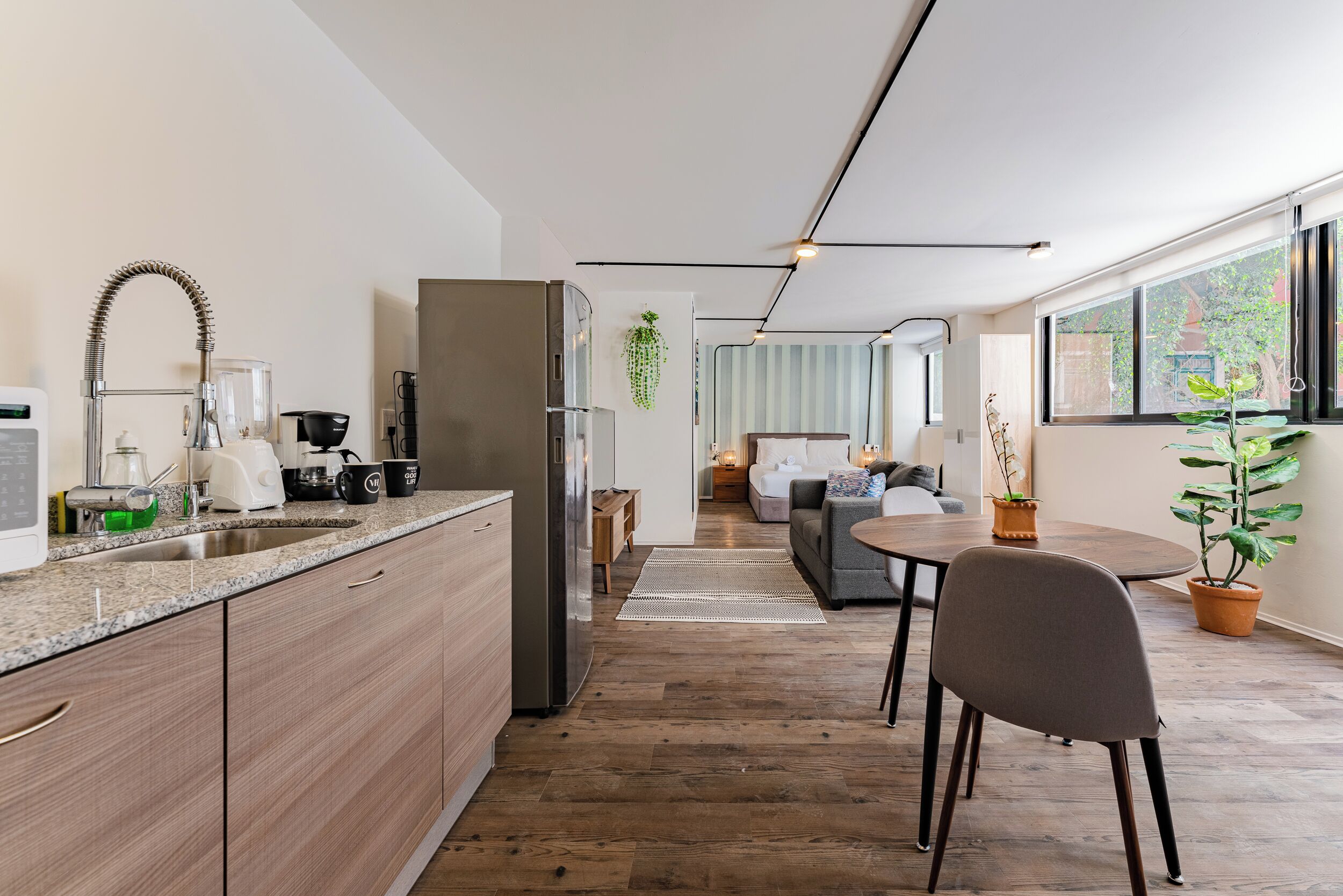 Una cocina moderna con armarios de madera, un refrigerador de acero inoxidable y un lavamanos. Hay una zona de comedor con una mesa y sillas, y una sala de estar con un sofá y una mesita de centro.