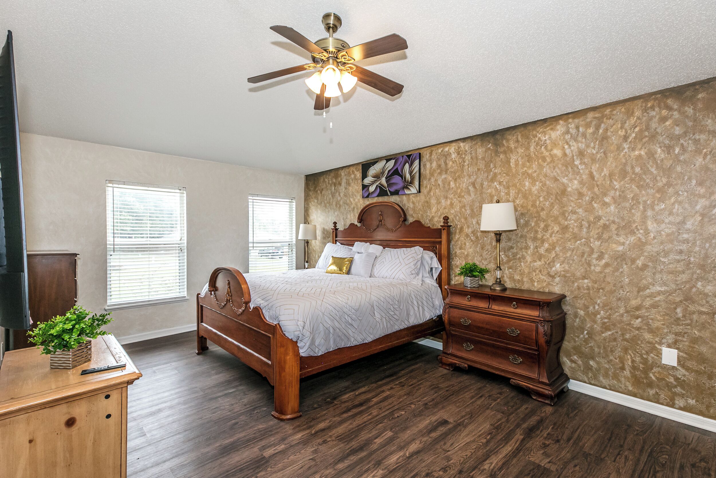 Une chambre à coucher avec un lit en bois, une table de chevet, un ventilateur de plafond et un mur orné d’un tableau représentant des motifs floraux.
