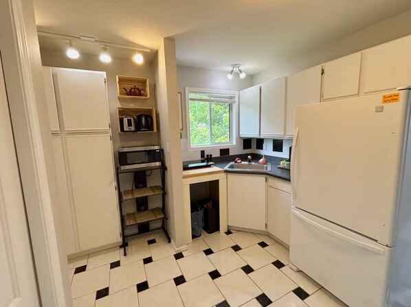 A compact kitchen with white cabinets, a microwave, a refrigerator, and a window providing natural light. - Hall's Haven: Simple & Cozy Escape. Pets Welcome! Hiking & Wine Trail Nearby. (West Kelowna)