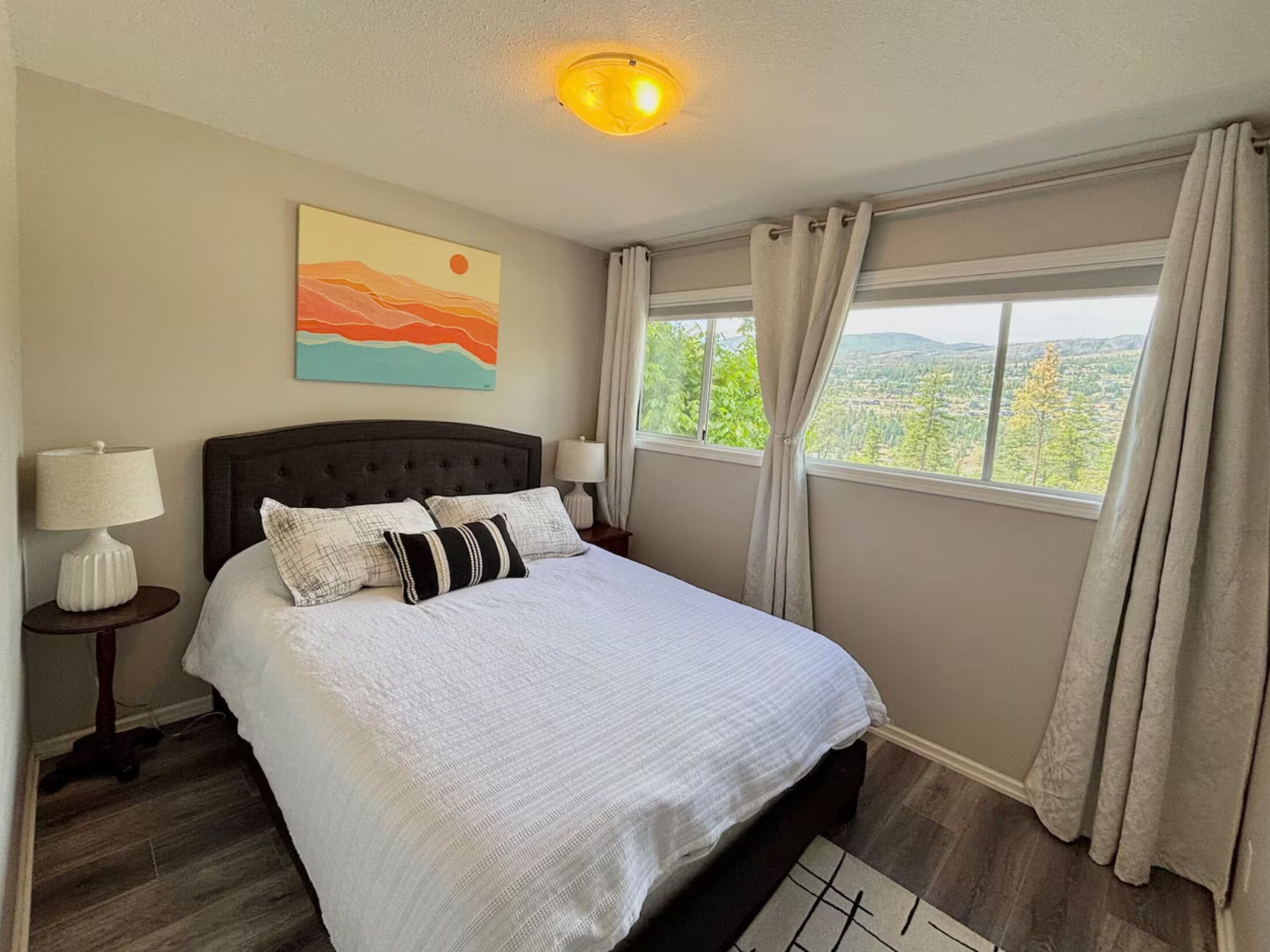 A bedroom with a bed, a nightstand, a lamp, a painting, and a window with curtains.