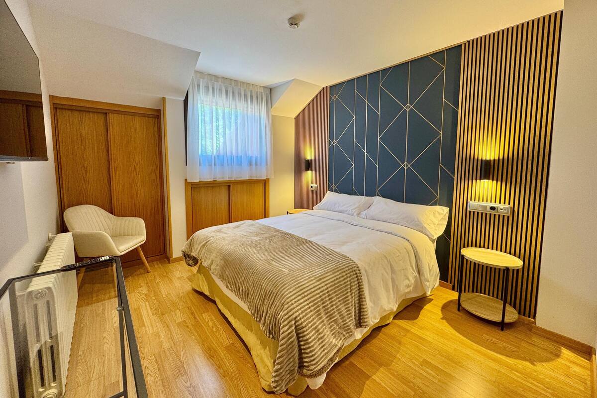 A modern hotel room with a bed, a chair, a small table, and a window with curtains.