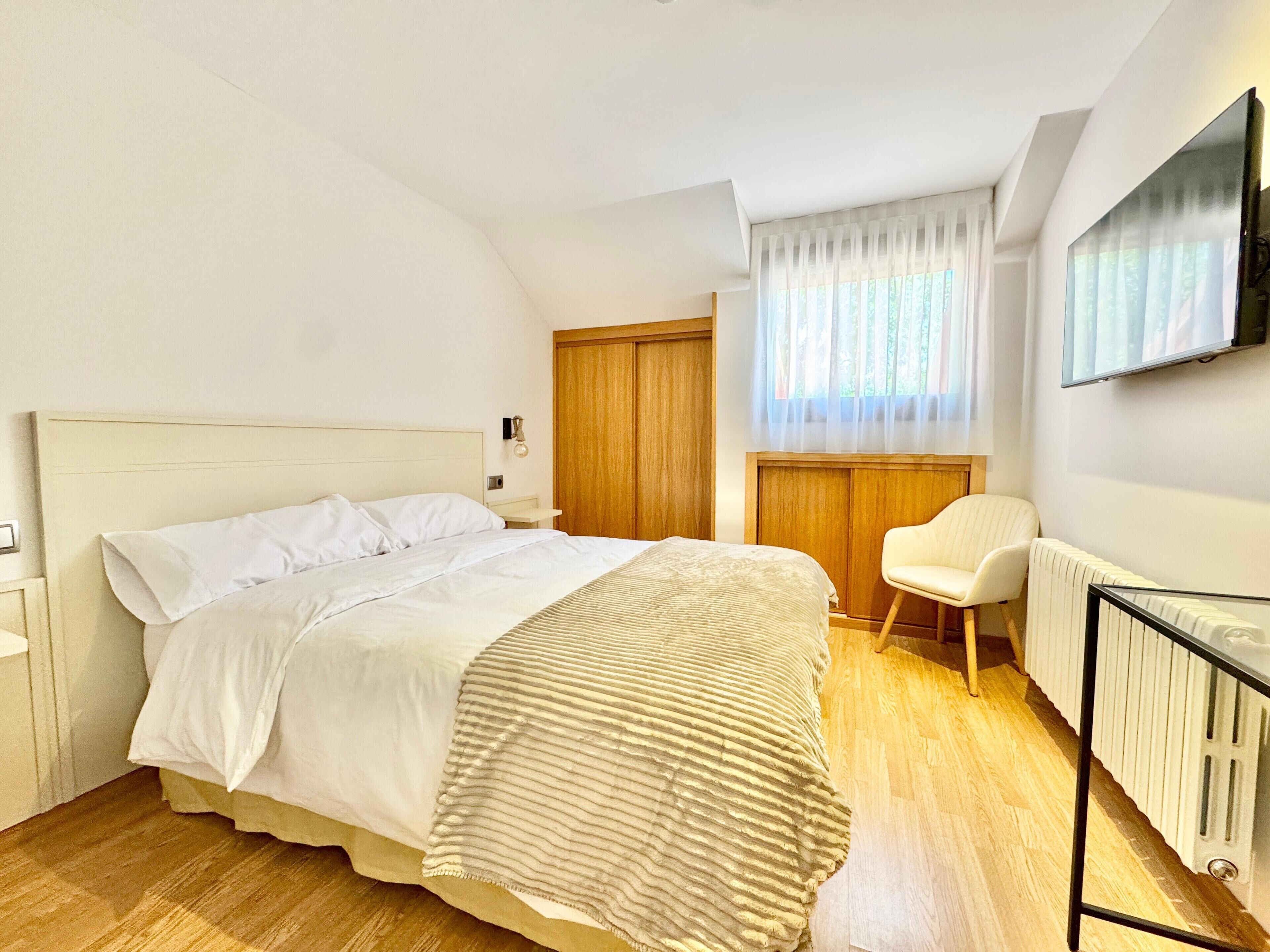 Un dormitorio con cama, una silla, puerta de madera, radiador y televisor montado en la pared.
