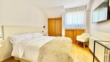 Un dormitorio con cama, una silla, puerta de madera, radiador y televisor montado en la pared.