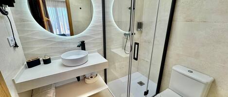 Un baño moderno con un espejo redondo, un lavabo blanco, ducha envuelta en vidrio y un inodoro.