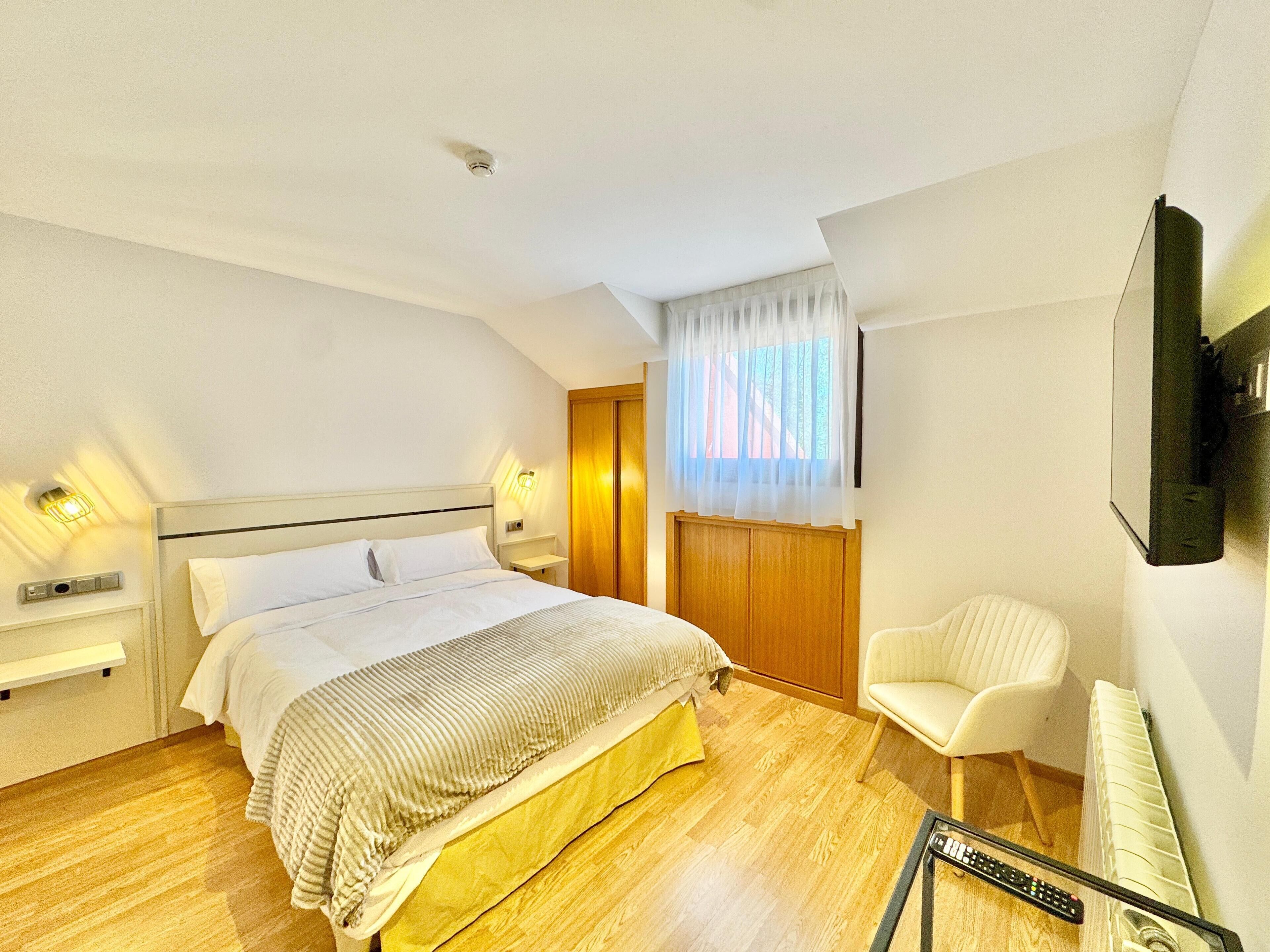 A bedroom with a bed, bedside table, lamp, chair, and a TV mounted on the wall.