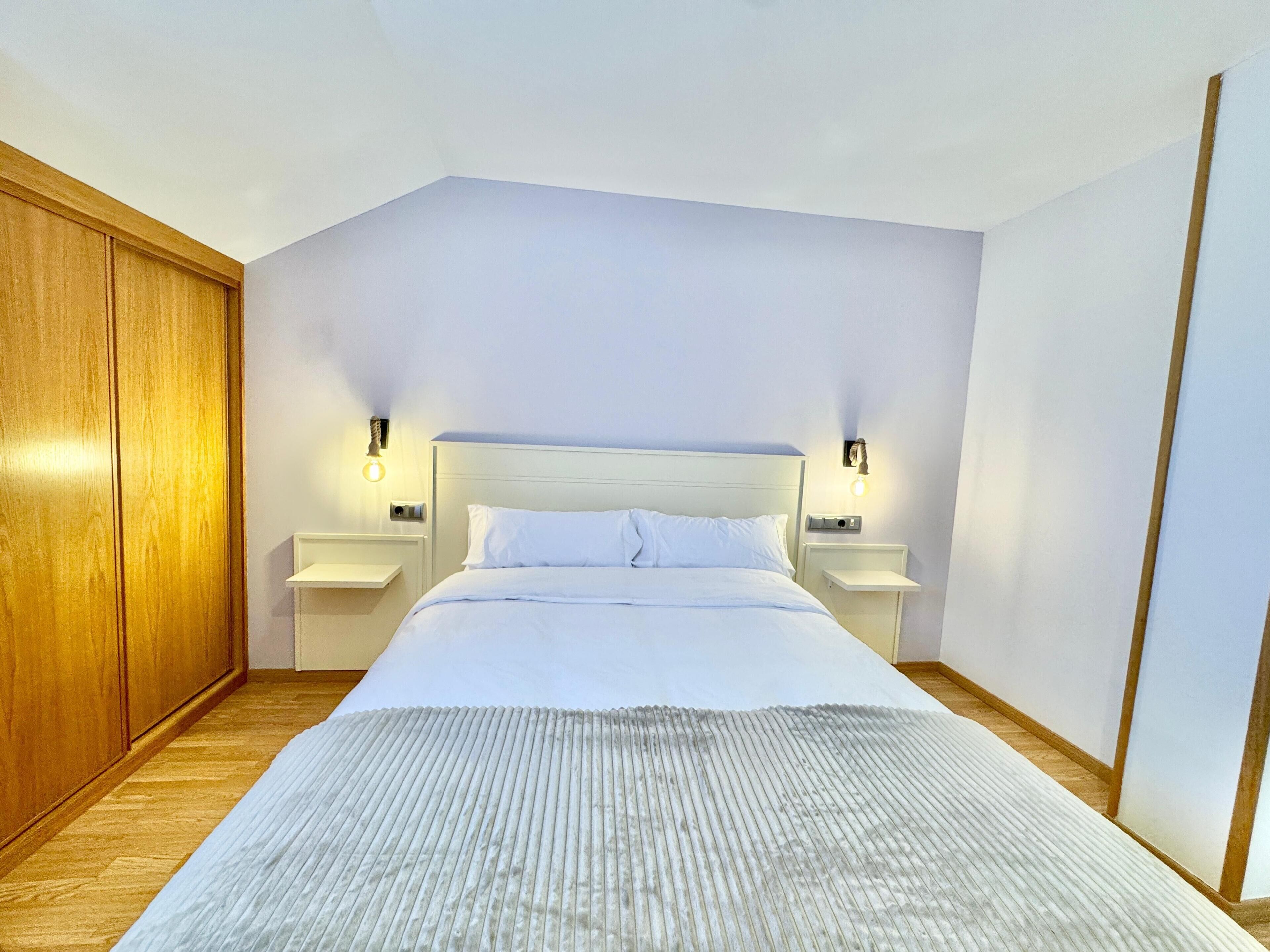 A bedroom with a large bed, wooden wardrobe, and two bedside tables with wall-mounted lamps.