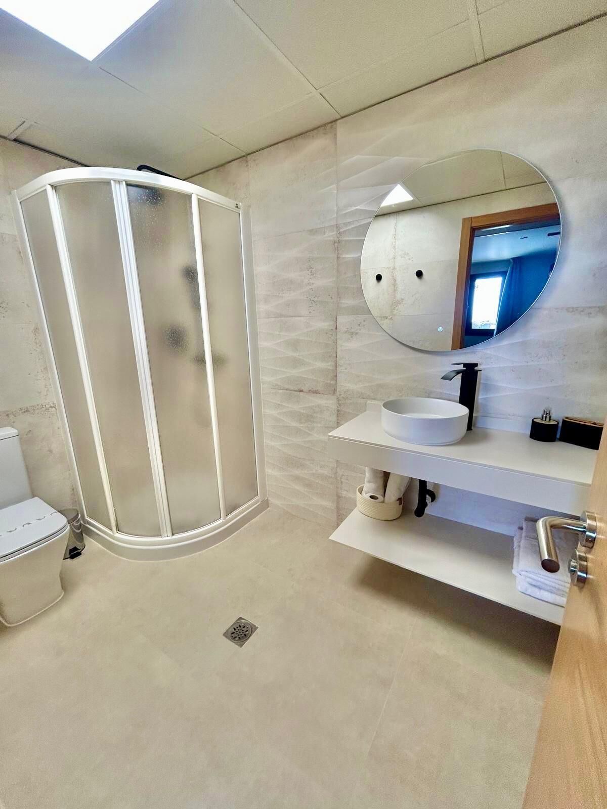 A modern bathroom with a glass-enclosed shower, a round mirror, and a white sink.