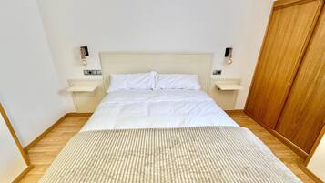 A neatly made bed with white linens in a room with wooden flooring and a wooden wardrobe.
