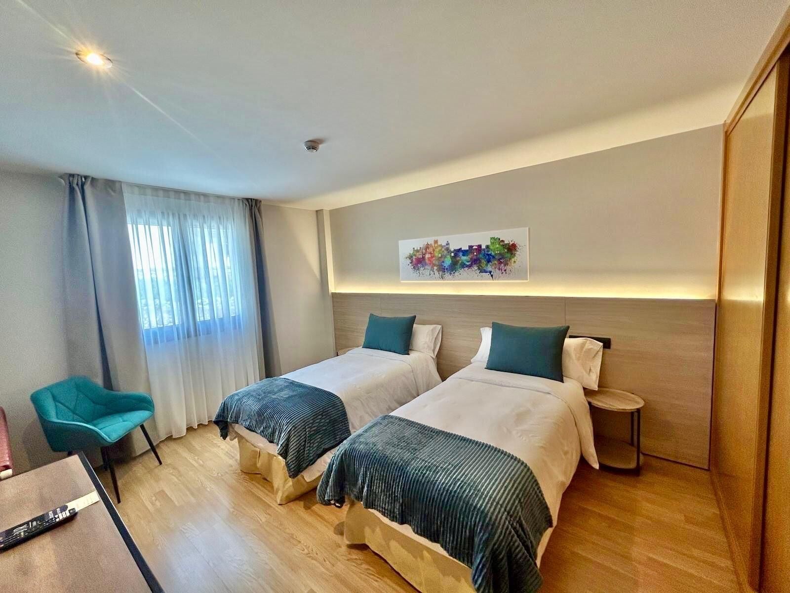 A hotel room with two beds, a chair, a small table, and a painting on the wall.