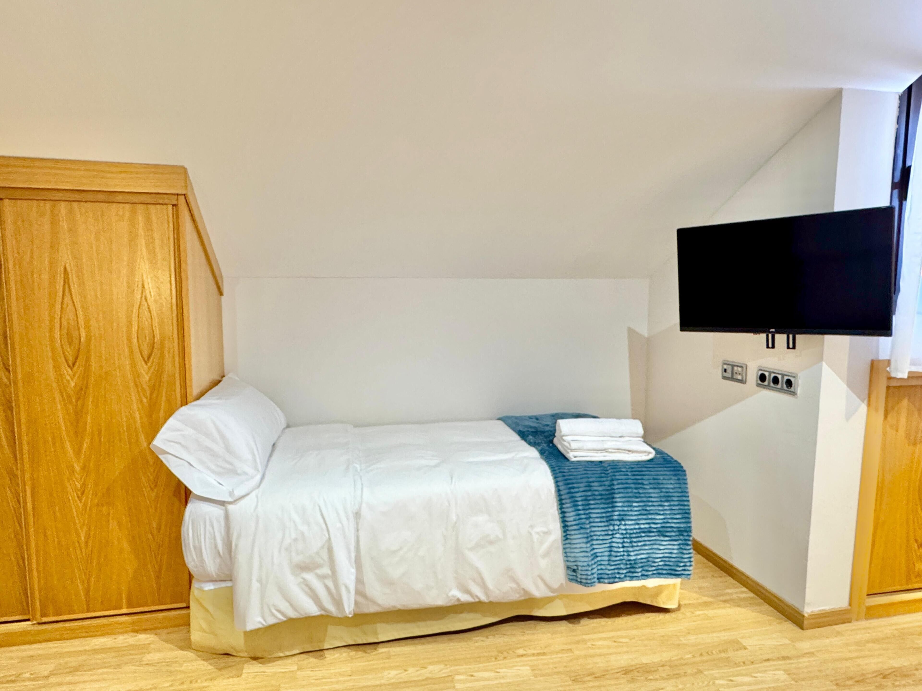 Una habitación pequeña con cama individual, un armario de madera, un televisor de pantalla plana montado en la pared y suelo de madera.