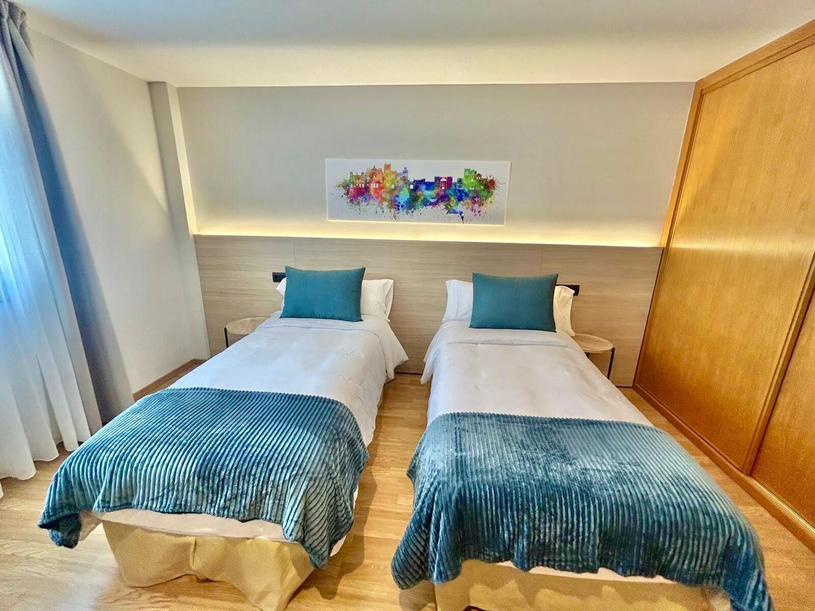 Une chambre d’hôtel avec deux lits, chacun recouvert d’une courtepointe bleue et blanche, un plancher en bois et une peinture abstraite aux couleurs vives accrochée au mur.