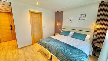 Un dormitorio con suelo de madera, una cama con edredón azul y blanco, una puerta de madera y una lámpara fijada a la pared.
