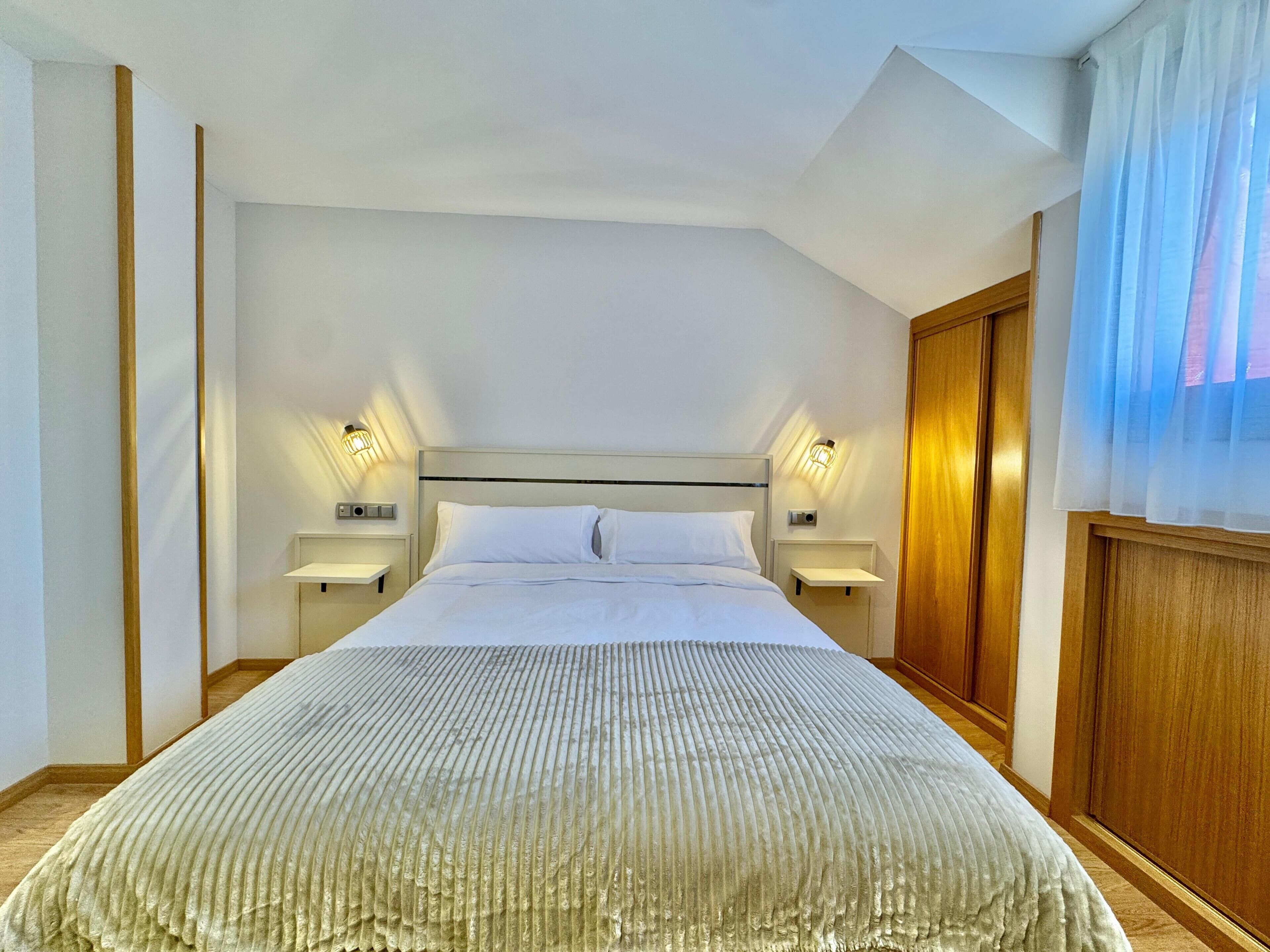 Quarto de hotel com uma cama grande, duas mesas de cabeceira com abajures, um guarda-roupa de madeira e uma janela com cortinas finas.