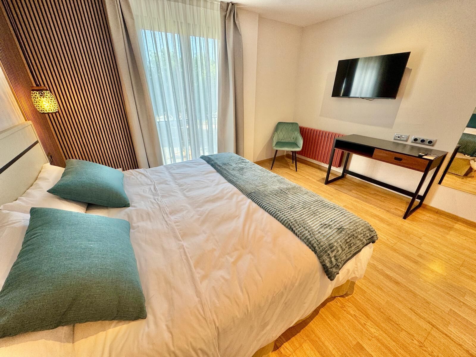 A modern hotel room with a large bed, a desk, a TV, and a window with curtains.