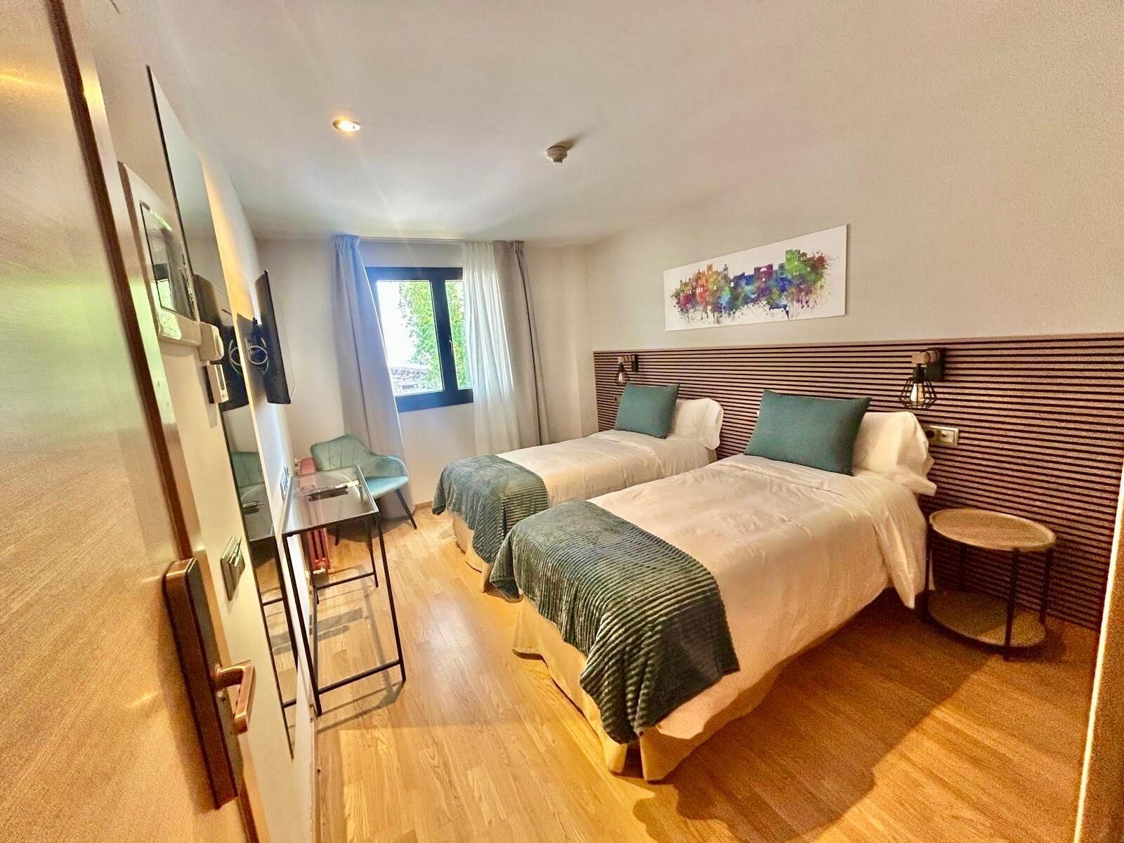 Ein Hotelzimmer mit zwei Betten, einem Schreibtisch, einem Stuhl, einem kleinen Tisch und einem Fenster mit Vorhängen.