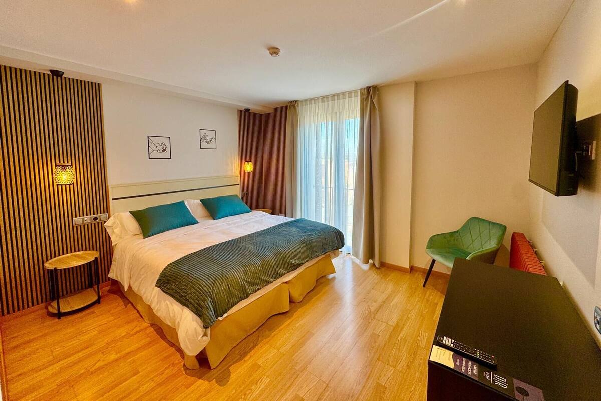 A hotel room with a large bed, a small table, a chair, a TV, and a window with curtains.
