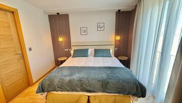 A modern hotel room with a large bed, two bedside tables, a small wall-mounted shelf, and a window with curtains.