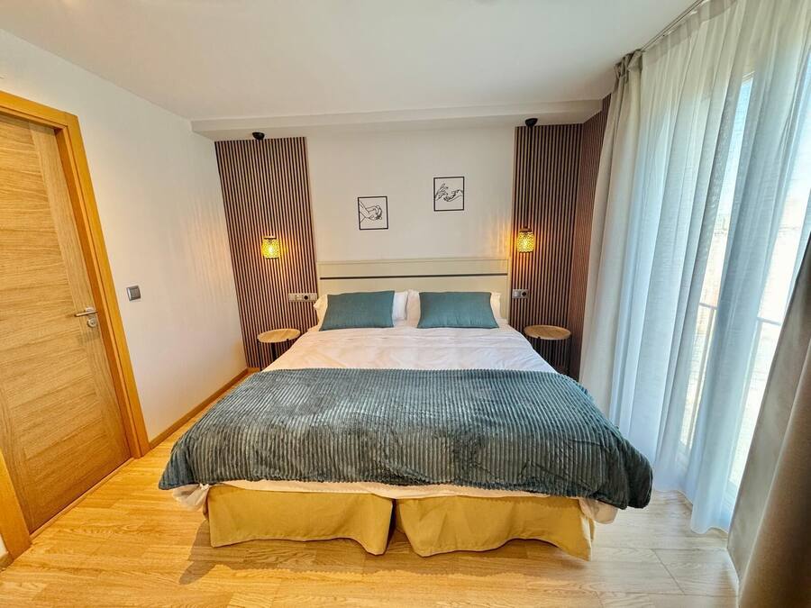 A modern hotel room with a large bed, two bedside tables, a small wall-mounted shelf, and a window with curtains.