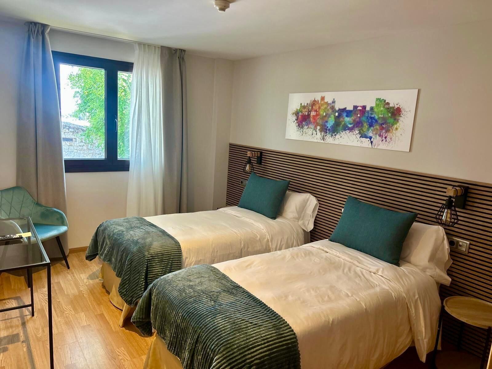A hotel room with two beds, a window with curtains, a chair, a small table, and a colorful abstract painting on the wall.