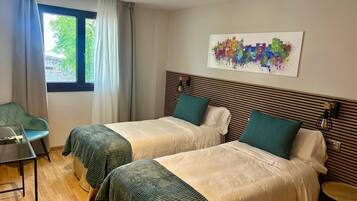 A hotel room with two beds, a window with curtains, a chair, a small table, and a colorful abstract painting on the wall.