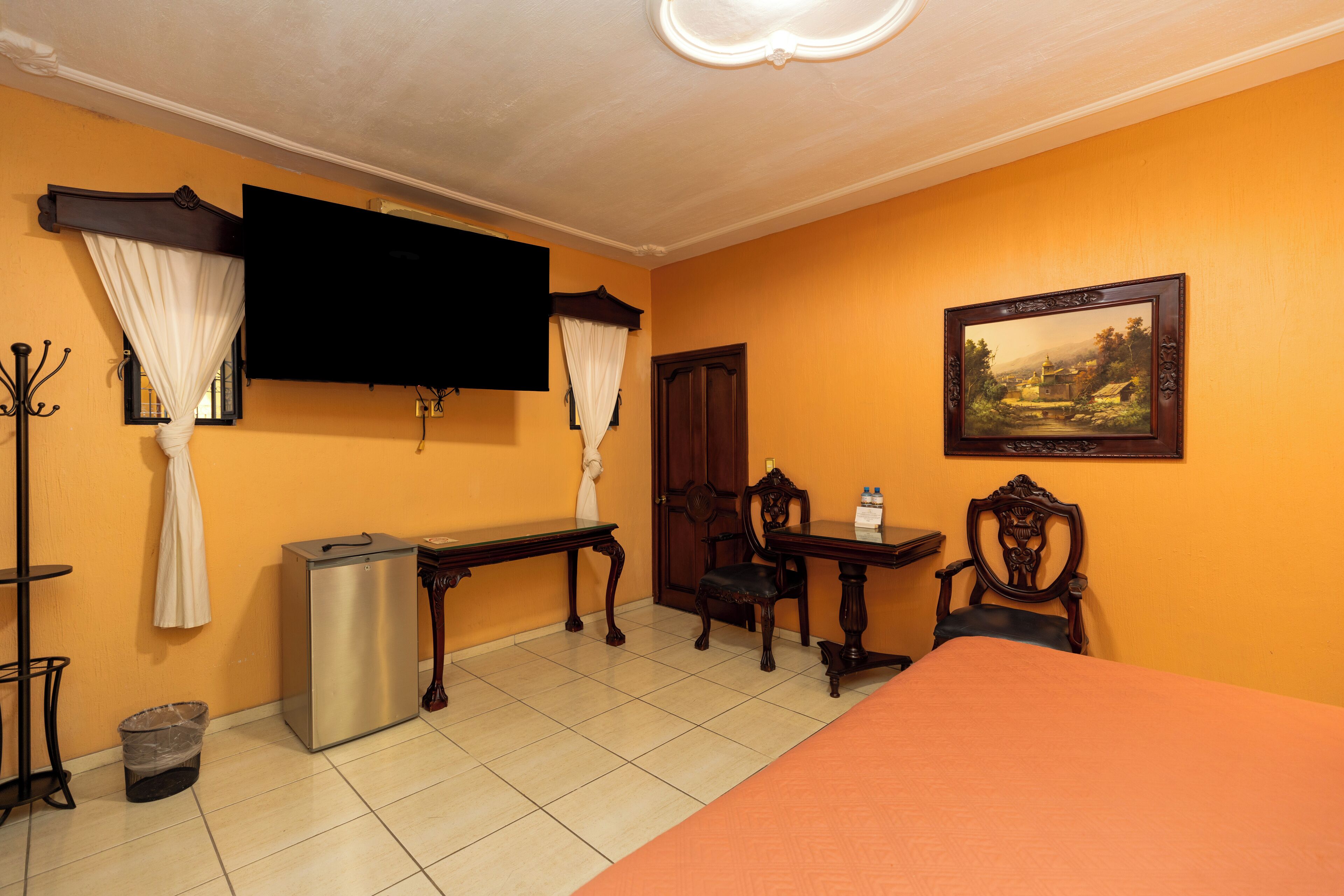 Quarto de hotel com cama, escrivaninha, cadeira, televisão e um quadro na parede.
