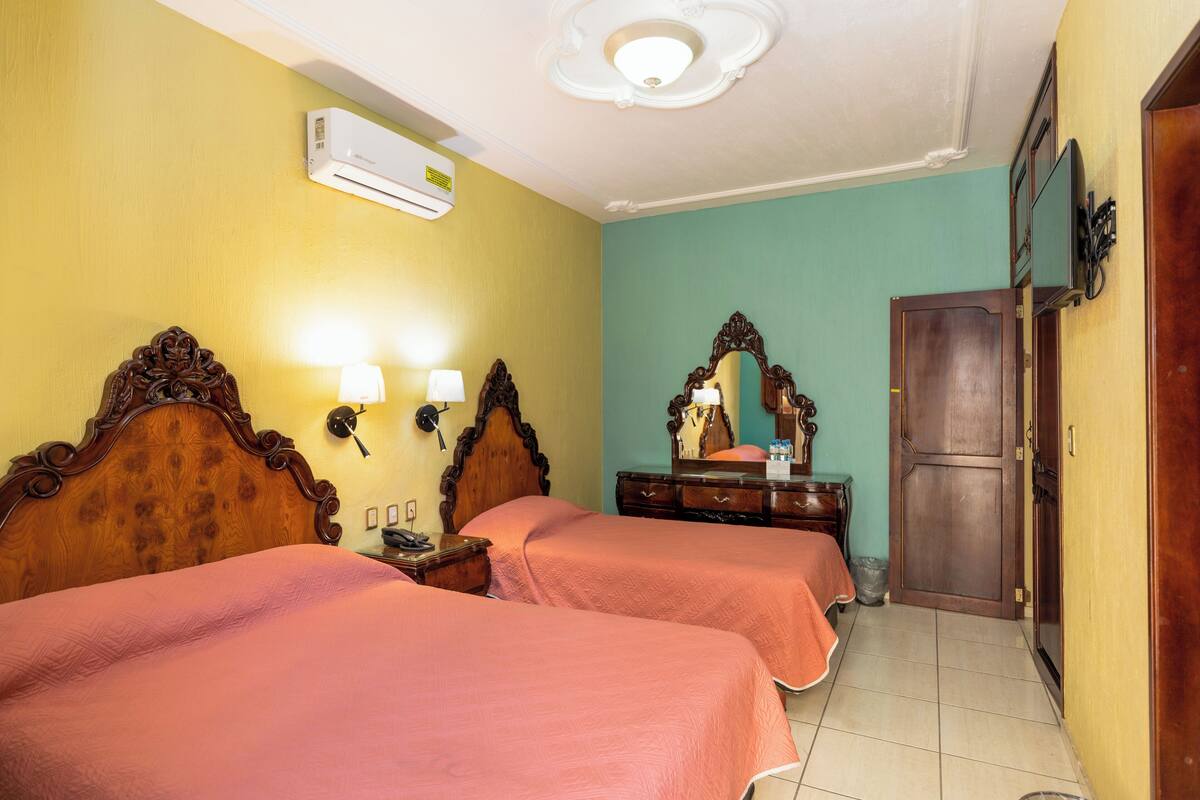 Une chambre d’hôtel avec deux lits, une tête de lit en bois, un miroir, une commode et une porte.