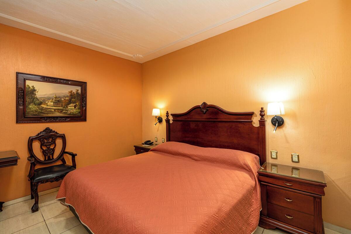 Une chambre d’hôtel avec un grand lit, une tête de lit en bois, deux lampes de chevet, une table de chevet, une chaise en bois et un tableau encadré accroché au mur.
