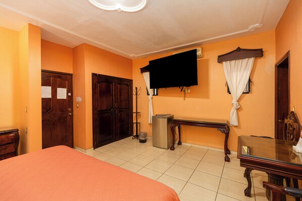 Una habitación de hotel con cama, escritorio, televisión, cesto de basura y ventana con cortinas.