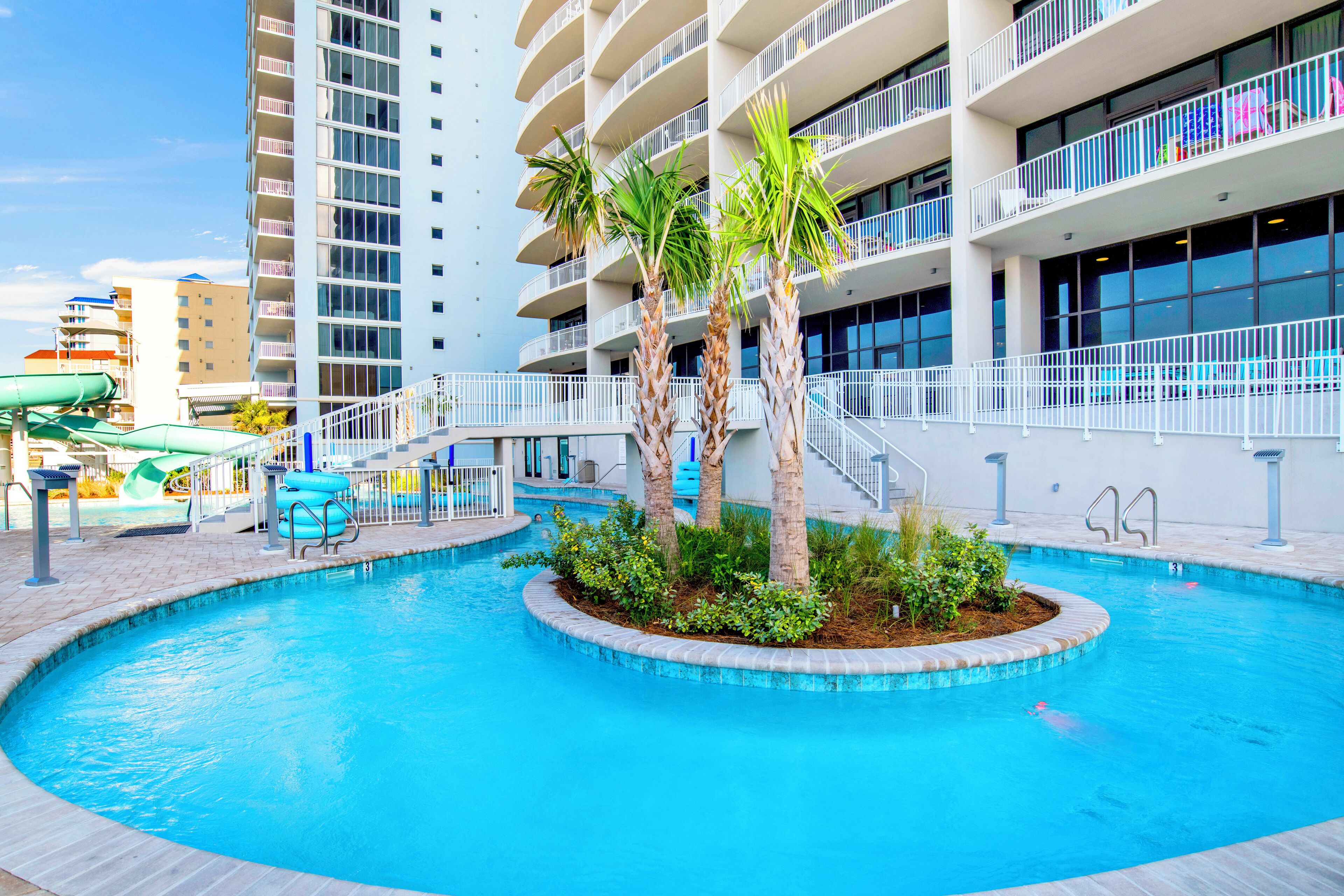 A swimming pool with palm trees, a water slide, and a multi-story building with balconies.