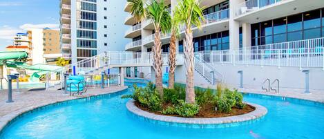 A swimming pool with palm trees, a water slide, and a multi-story building with balconies.