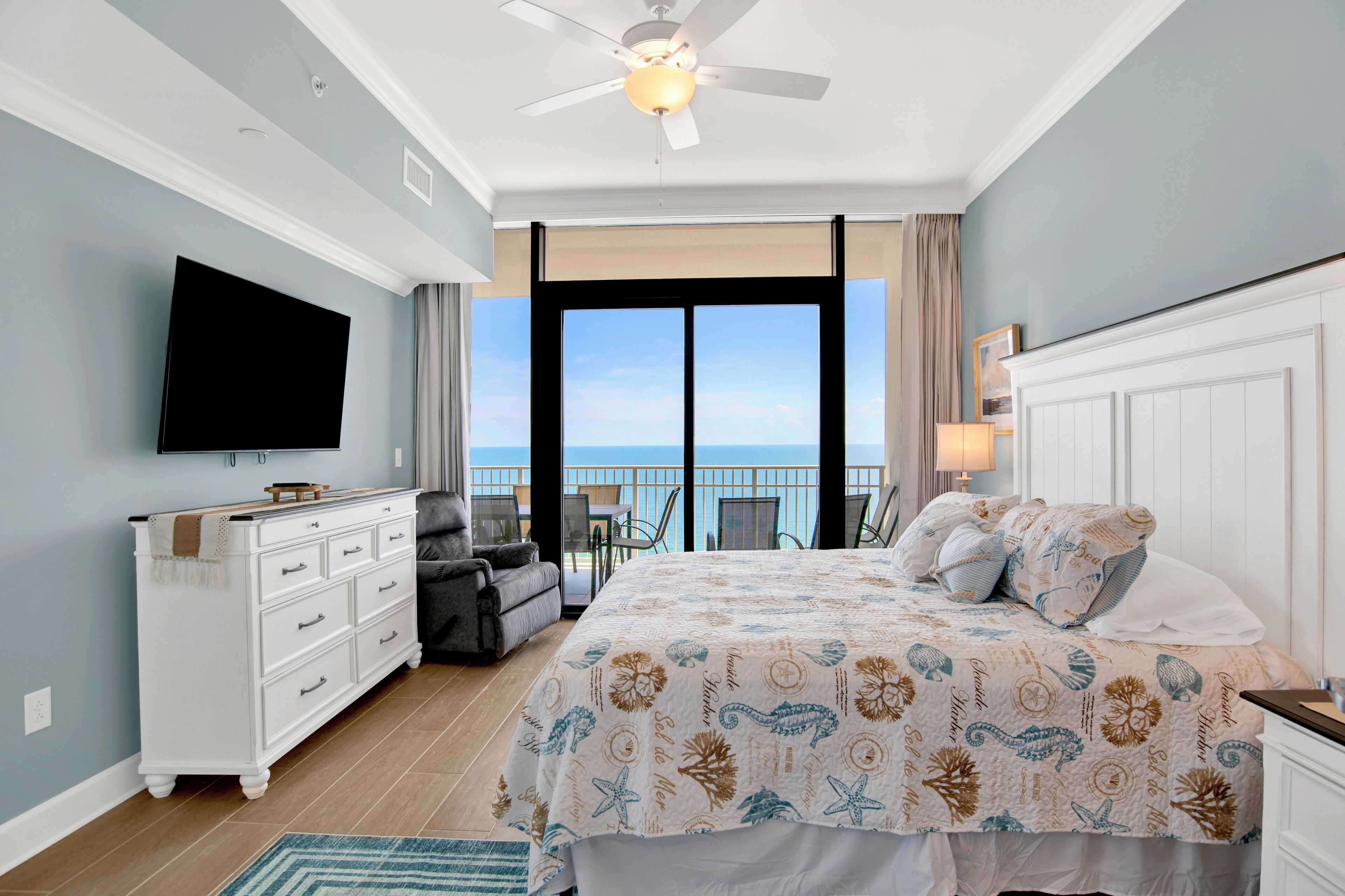A bedroom with a bed, a TV, a dresser, and a balcony with a view of the ocean.