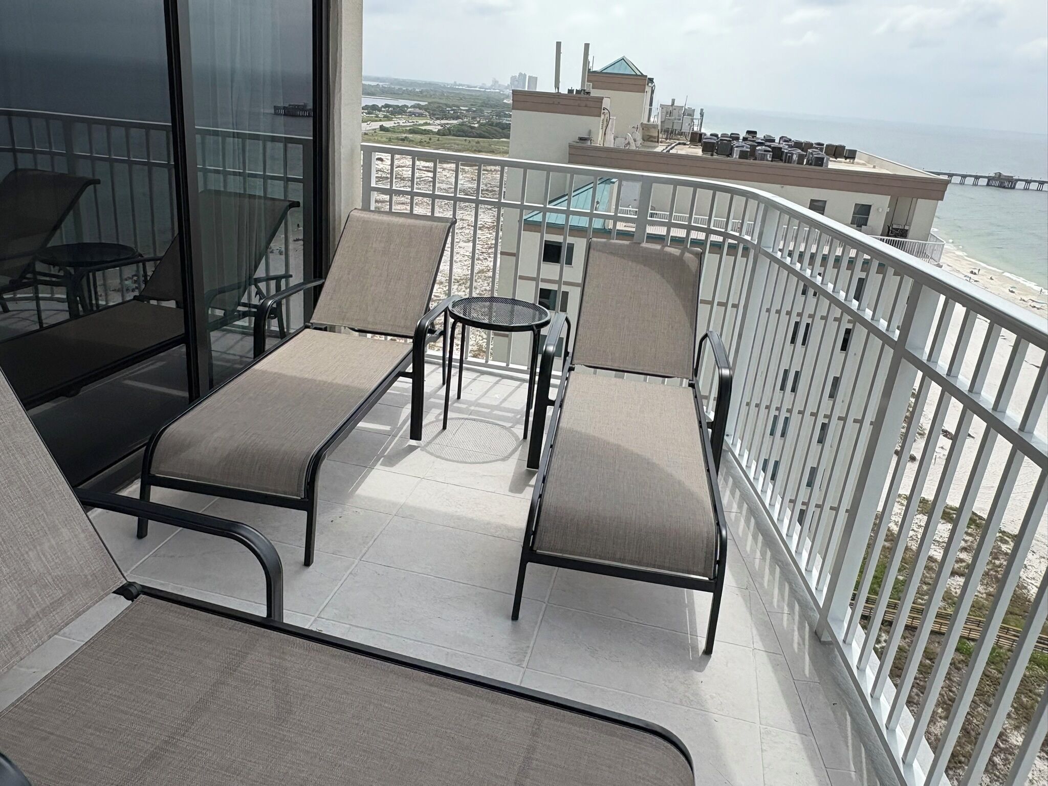 A balcony with two lounge chairs and a small table, overlooking a beach and cityscape.