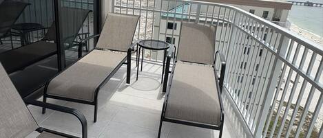 A balcony with two lounge chairs and a small table, overlooking a beach and cityscape.
