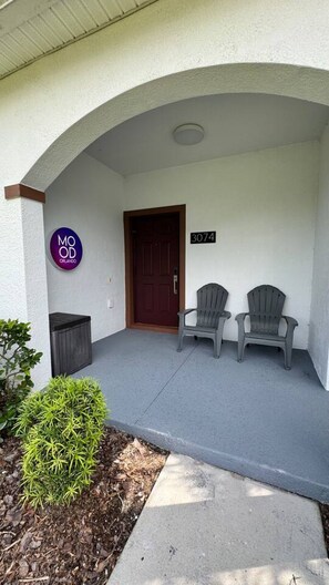 A building entrance with a covered porch, a door, two Adirondack chairs, a small bush, and a sign with 'MOOD ORLANDO'.